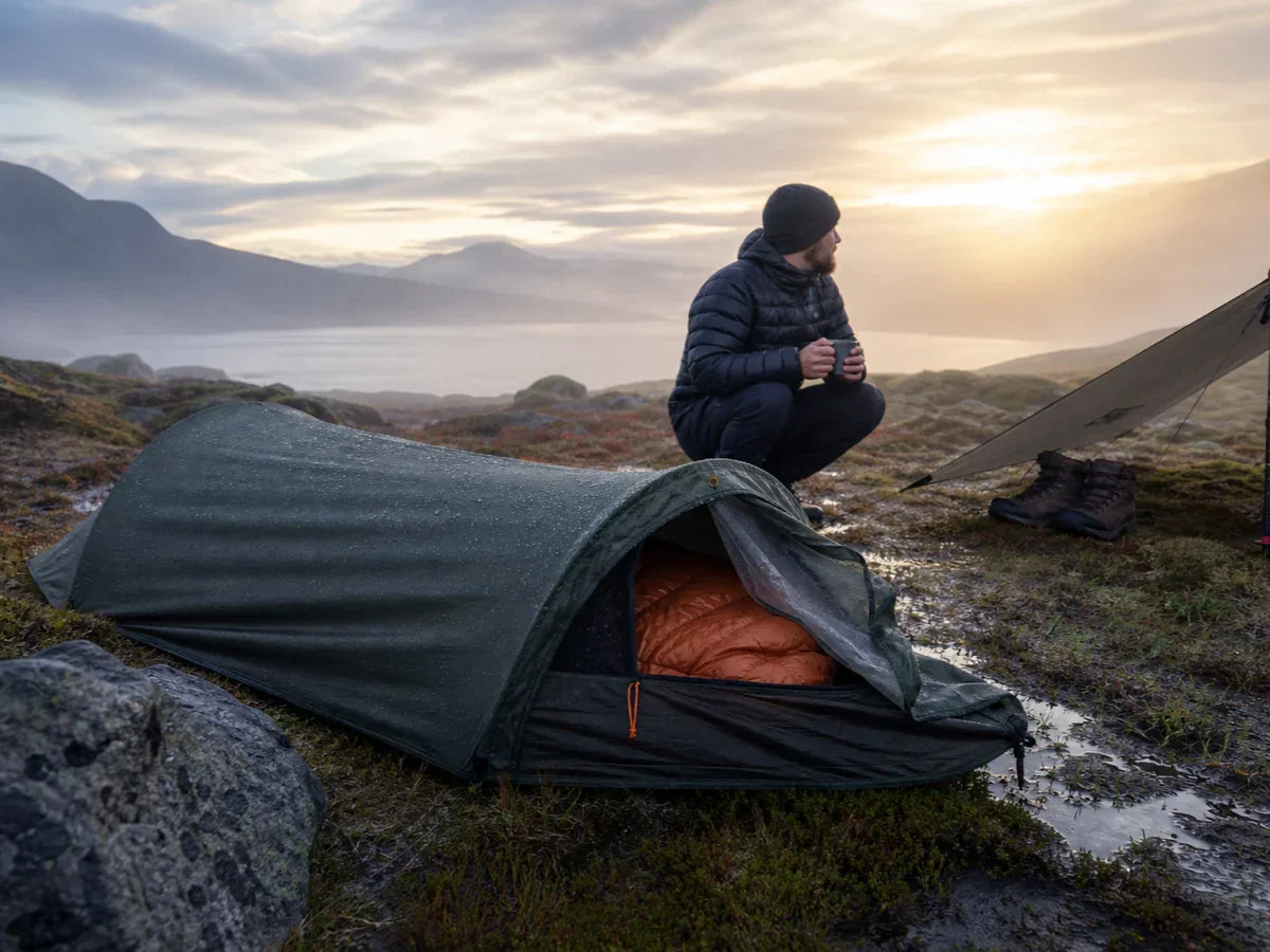 Solo camper beside a waterproof bivy-style shelter after rain at sunrise.